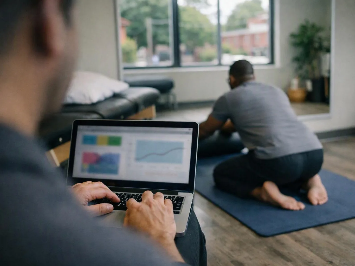 A person types on a laptop while another kneels on a yoga mat in a bright studio room