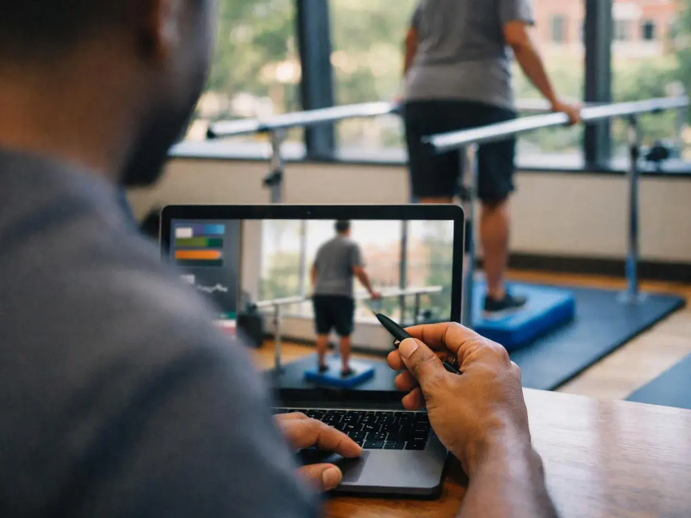 A person uses a laptop to watch someone balance on a blue platform in a bright exercise room